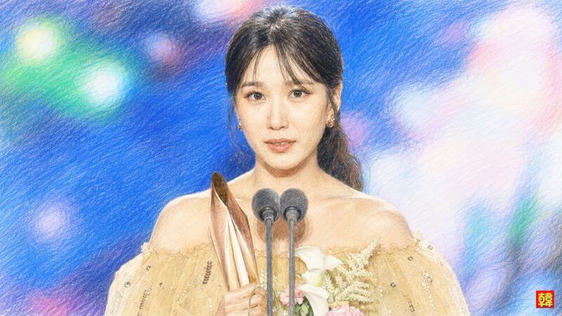 華やかな授賞式のステージに立つパク・ウンビン。オフショルダーのゴールドのドレスを身にまとい、マイクの前でトロフィーと花束を抱えて微笑む、百想芸術大賞での感動的な受賞シーンを描いたイラスト。